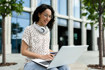 Smiling young woman with headphones using laptop while sitting on a bench outside, surrounded by modern urban architecture.