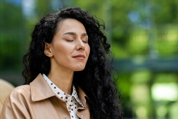 Serene young woman with eyes closed, savoring peace in a natural setting with blurred greenery in...