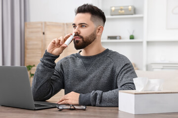 Medical drops. Young man using nasal spray at table indoors