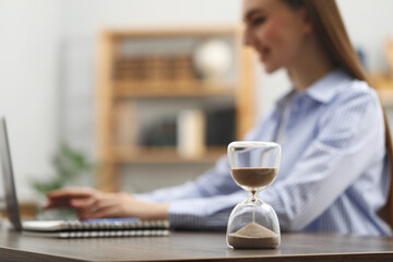 Hourglass with flowing sand on desk. Woman using laptop indoors, selective focus