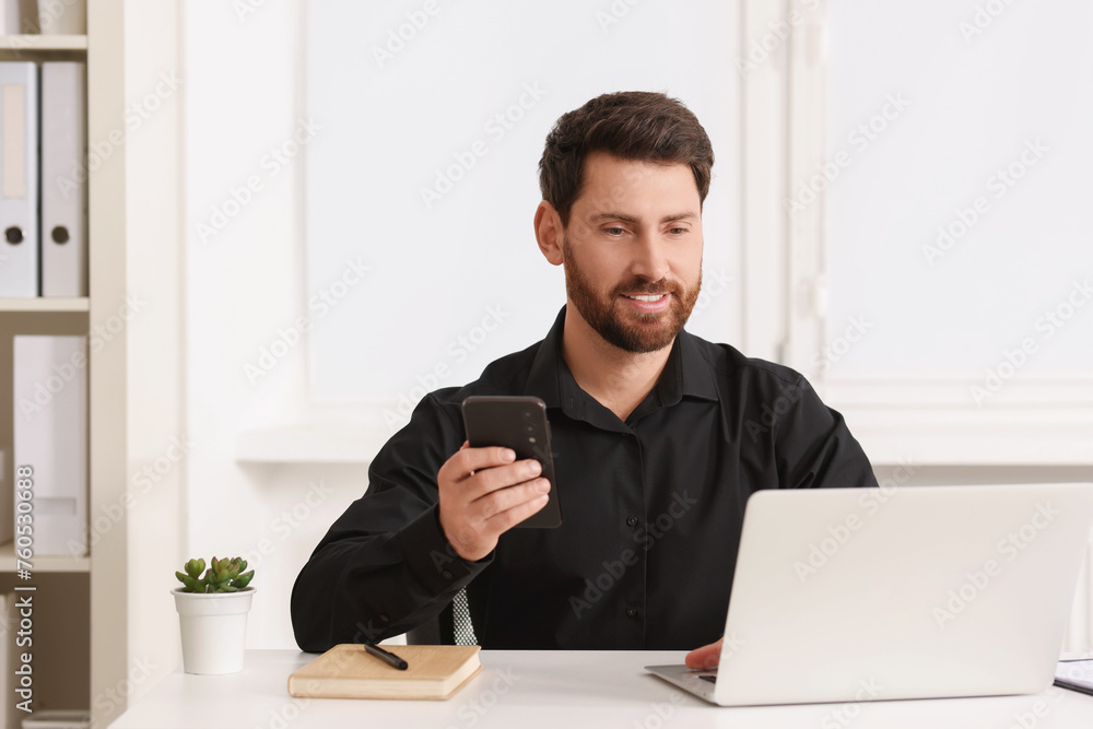 Poster smiling man with smartphone using laptop at table in office