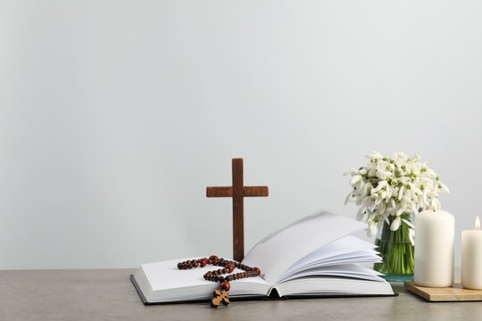 Church candles, Bible, wooden cross, rosary beads and flowers on grey table. Space for text