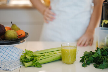 Close-up of detox drink in glass. Celery and parsley on the table. Background of female in apron. Healthy homemade beverage made in juicer