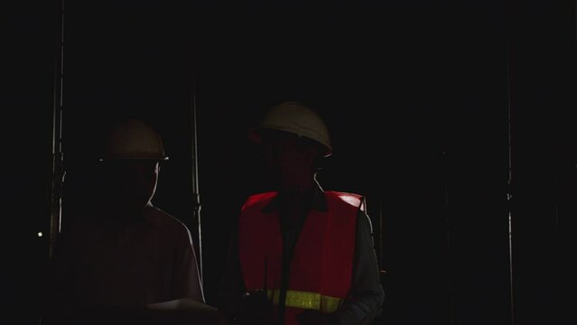 Working Late at Night in construction site. Engineers and architects in safety uniforms and hard hats talk and review blueprints as they  walking in a building site.