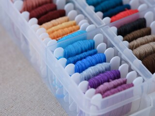 A close-up shot of a plastic sorting box full of bobbins with different colour embroidery threads on a beige canvas background.