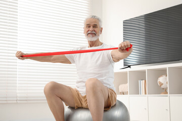 Fototapeta na wymiar Senior man doing exercise with elastic resistance band on fitness ball at home