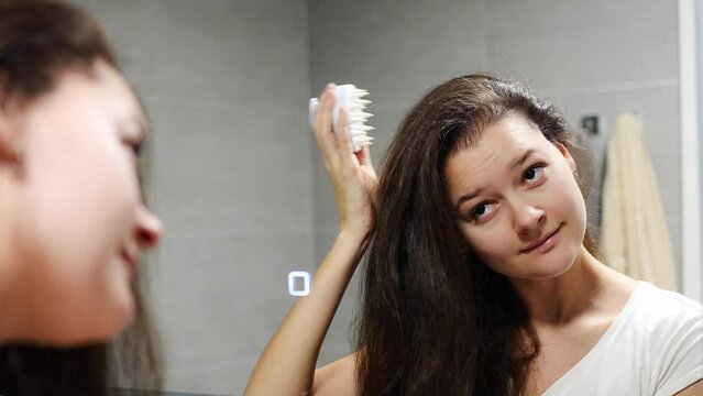 Young woman with dark curly hair doing self hair scalp massage with scalp massager or hair brush for hair growth stimulating at home bathroom. Reflected view of the mirror