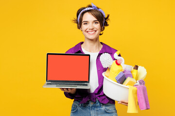 Young IT woman wear purple shirt hold basin with detergent bottles do housework tidy up work use blank empty area screen laptop pc computer isolated on plain yellow background. Housekeeping concept.