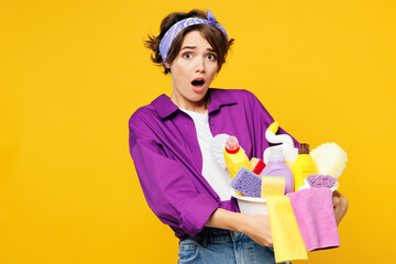 Young shocked woman wear purple shirt hold basin with detergent bottles do housework tidy up looking camera with opened mouth isolated on plain yellow background studio portrait. Housekeeping concept.