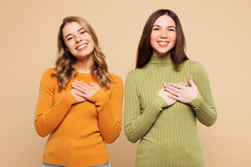 Young grateful smiling friends two women wear orange green shirt casual clothes together put folded hands on heart isolated on plain pastel light beige background studio portrait. Lifestyle concept.