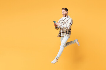 Full body side view young Caucasian man he wear brown shirt casual clothes jump high hold in hand use mobile cell phone isolated on plain yellow orange background studio portrait. Lifestyle concept.