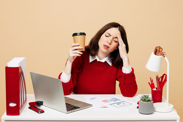 Young tired employee business woman wear red sweater shirt sit work at office desk with pc laptop hold takeaway delivery cup coffee to go isolated on plain beige background Achievement career concept