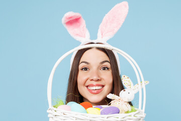 Close up cropped young woman wear pink casual clothes rabbit bunny ears look camera through wicker basket with eggs isolated on plain blue background studio portrait. Lifestyle Happy Easter concept.
