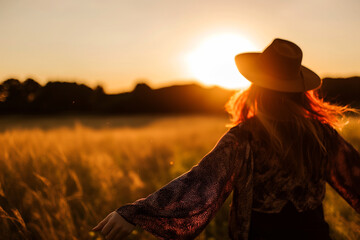 A woman in a hat stands in a field of tall grass at sunset - Powered by Adobe