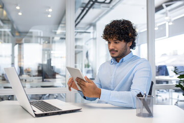 A man dressed in a blue shirt engages with a tablet at his orderly workstation in a bright,...
