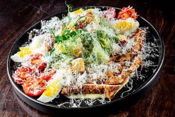 fresh Caesar salad with grilled chicken, parmesan, eggs, and tomatoes on a dark plate set against a wooden background. The vibrant green lettuce contrasts with the browned chicken