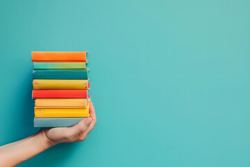 Hand holding a stack of colorful books isolated on a blue background, in a minimalistic style, with copy space for text - obrazy, fototapety, plakaty