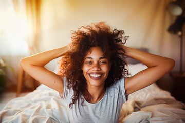 A joyful young woman with curly hair smiling brightly in a sunny bedroom, exuding happiness and comfort. - obrazy, fototapety, plakaty