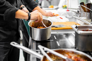 busy professional kitchen where a chef, dressed in dark attire, stirs a stainless steel pot. Cooking utensils, ingredients, and other pots are visible. The scene exudes culinary energy and creativity
