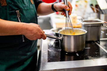 person in a green apron vigorously whisking a mixture in a stainless steel pot on a stove in a professional kitchen. The surroundings are blurred, focusing on the cooking activity