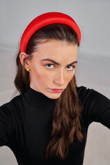 Close-up shot of a young beautiful woman girl with loose hair wearing a black turtleneck and a red headband. European girl in a fashionable wide headband taking selfies against a gray background.