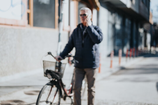 Blurred image of a senior adult with a bike on an urban road, engaged in a call on his mobile phone. Captures the essence of active city life and connectivity.