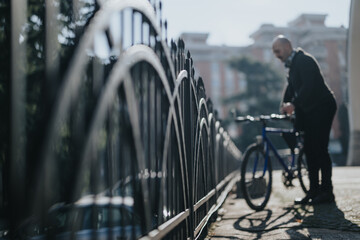 A bald man in casual attire stands beside his bicycle on a sunny urban street, with a shallow depth...