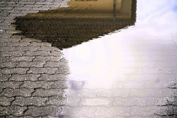 Große Regenpfütze auf grauem Backsteinboden mit Spiegelung von gelbem Haus und Himmel bei Regen...