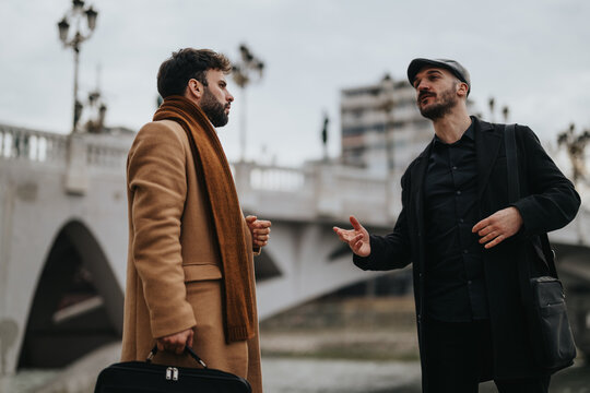 A focused outdoor shot capturing two men in professional attire having a serious discussion in a city setting with a bridge background.