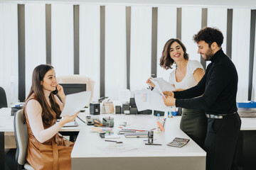 Three colleagues in a bright office, with one woman on a phone call and two laughing while looking at documents.