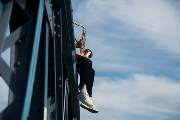 Closeup of musician playing saxophone sitting on a metallic bridge in the street - 760725218