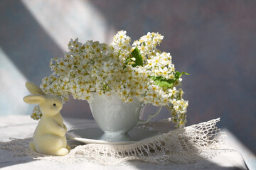 Spring bouquet with bird cherry flowers in a cup on the table, a figurine of a rabbit, sunlight, Easter still life. - 760727400