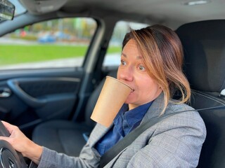 Close up of Caucasian businesswoman driving car in city looking at road with big opened eyes holding coffee in mouth. Young female drives automobile with paper cup of coffee