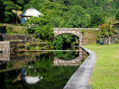 Vista de un paisaje con un puente de piedra al fondo con &aacute;rboles verdes reflejado en el agua del r&iacute;o y c&eacute;sped en la orilla derecha con bordillo de piedra, en Orense, verano de 2021