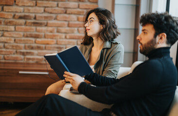 Two professionals in a casual office setting, woman reading a book with interest, man in contemplation, warm brick backdrop.