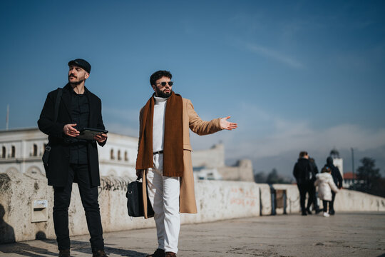 Urban scene featuring two fashionable men on a city street: one hailing a cab or asking for directions, the other holding a digital tablet.