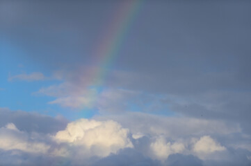 Fototapeta na wymiar Detail Regenbogen vor schwarzen und weißen Wolken und kleinen Flecken blauem Himmel