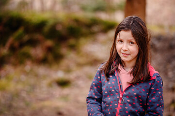 A cheerful young girl with brown hair smiles brightly, wearing a dotted jacket in a lush forest setting, exuding warmth and innocence