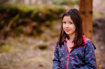 Fototapeta premium A cheerful young girl with brown hair smiles brightly, wearing a dotted jacket in a lush forest setting, exuding warmth and innocence
