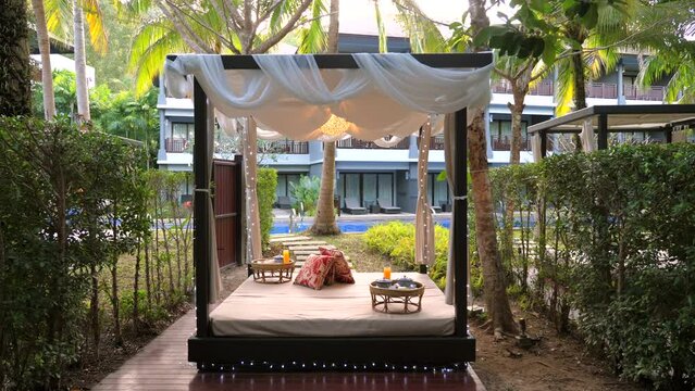 Luxurious outdoor hotel cabana draped with sheer white curtains and furnished with plush daybed, overlooking tranquil pool and lush greenery. Tropical resort relaxation and comfort.