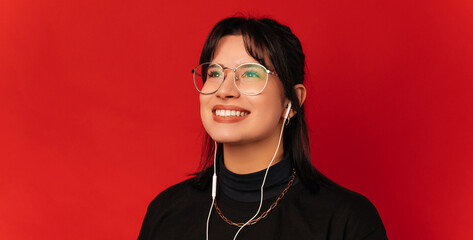 Studio shot of a young dreamy smiling woman wearing earphones over red background.