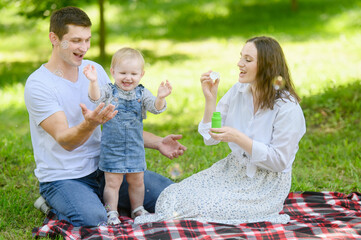 Happy family playing with soap bubbles during a picnic in nature on a warm sunny day, smiling, having fun, spending time together. Concept of happy, harmonious family relationships, family weekends.