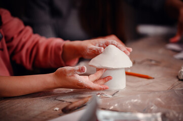 A child's hands carefully construct a clay house, with the focused guidance of a parent,...