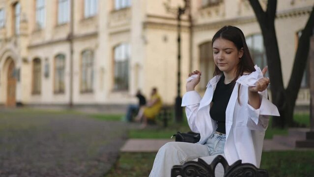 A girl sits on a bench with a notebook on her lap, in the institute park against the backdrop of a building. The girl rips a sheet out of the notebook, tears it up and throws it away