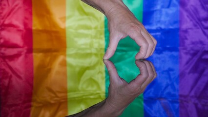 Male hand reflects on glass surface forming a heart with Gay and LGBT flag in the background