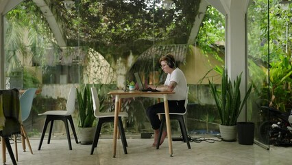 Transgender entrepreneur works late on project in serene, plant-filled open work space, typing diligently, focused on laptop screen. Pro stays overtime in peaceful, verdant coworking area, tasks.