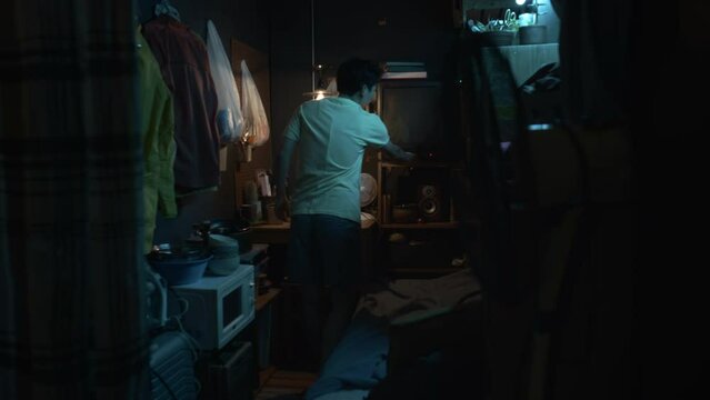 Full shot of young Asian man walking into micro apartment, switching on light bulb, turning on TV, laying down on bunk bed, pulling over blanket and watching movie, while resting at home after work