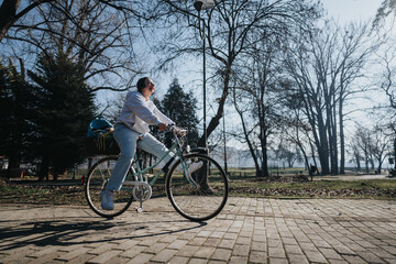 Fototapeta premium A joyful female cyclist with headphones on rides along a paved park path surrounded by bare trees and a clear sky.