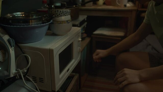 Medium close-up tilt shot of young Chinese man or woman sitting on bunk bed in tiny apartment, opening microwave oven, putting in food in plastic container and switching on for heating