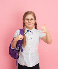 Smiling schoolgirl in eyeglasses with backpack showing thumb up gesture on pink background. Education and back to school concept.